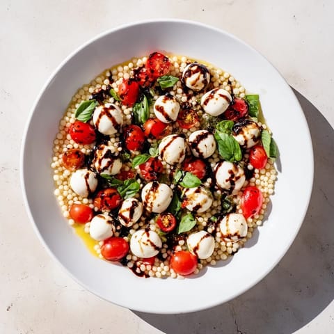Caprese Couscous Salad auf einem rustikalen Holztisch mit glänzenden Kirschtomaten, Mozzarella-Kugeln und frischen Basilikumblättern, übergossen mit Balsamico-Glasur.